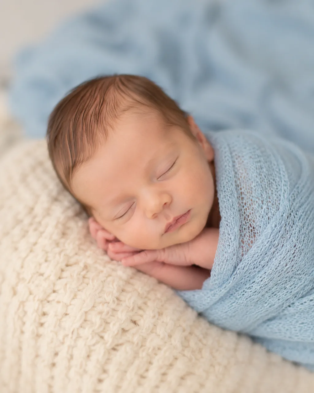 A peaceful sleeping newborn swaddled in a soft blue wrap, resting calmly on a cream blanket